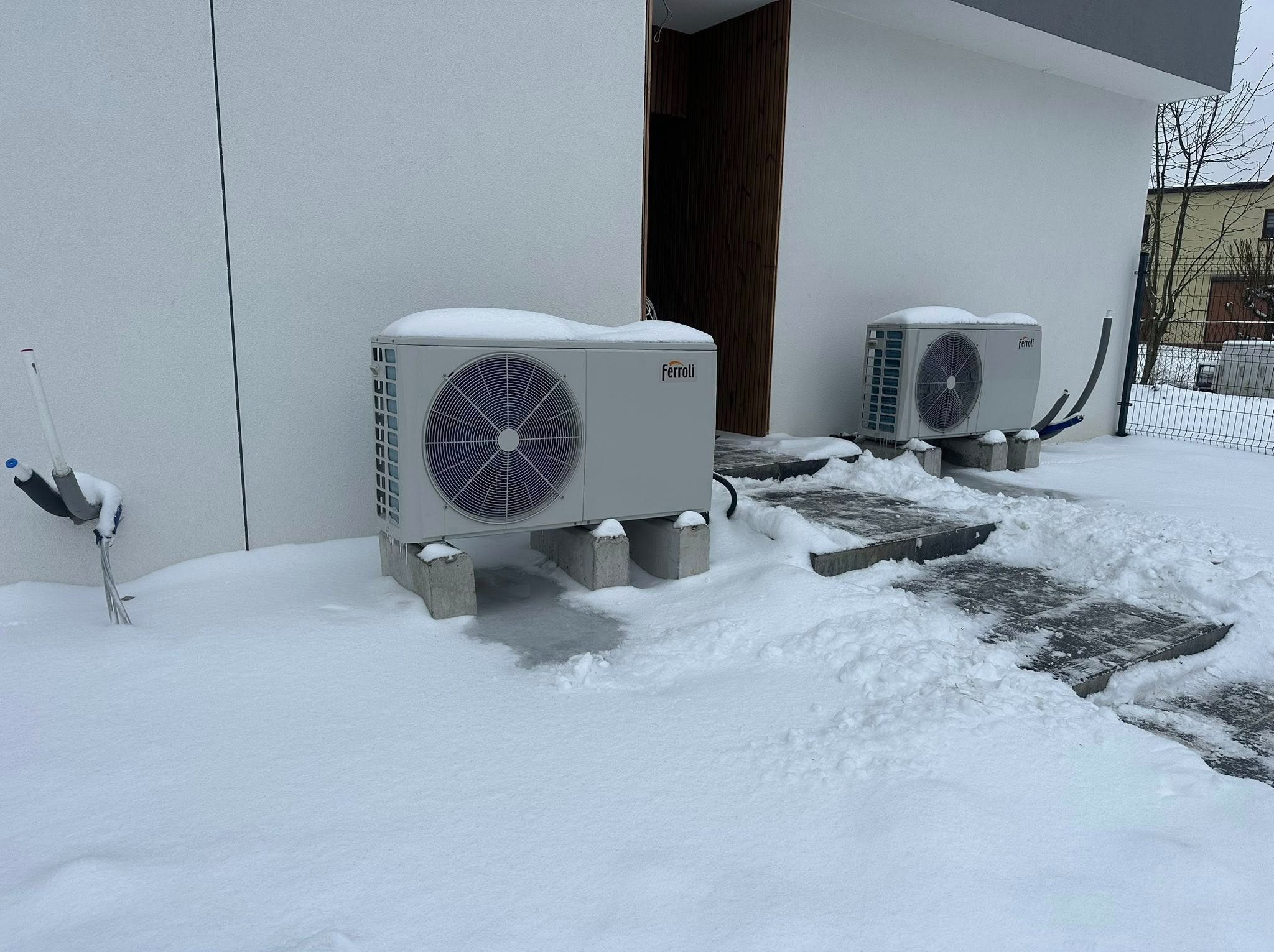Dwie pompy ciepła Ferroli, przysypane śniegiem, stoją na betonowych podstawach przed budynkiem w zimowej scenerii. Widoczne rury i elementy instalacji.