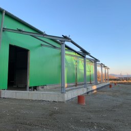 Olo-Car Aleksander Ostrowski - Zielona hala w trakcie budowy, widoczne stalowe elementy konstrukcyjne i fundamenty. W tle krajobraz z drzewami i wzgórzami, błękitne niebo.