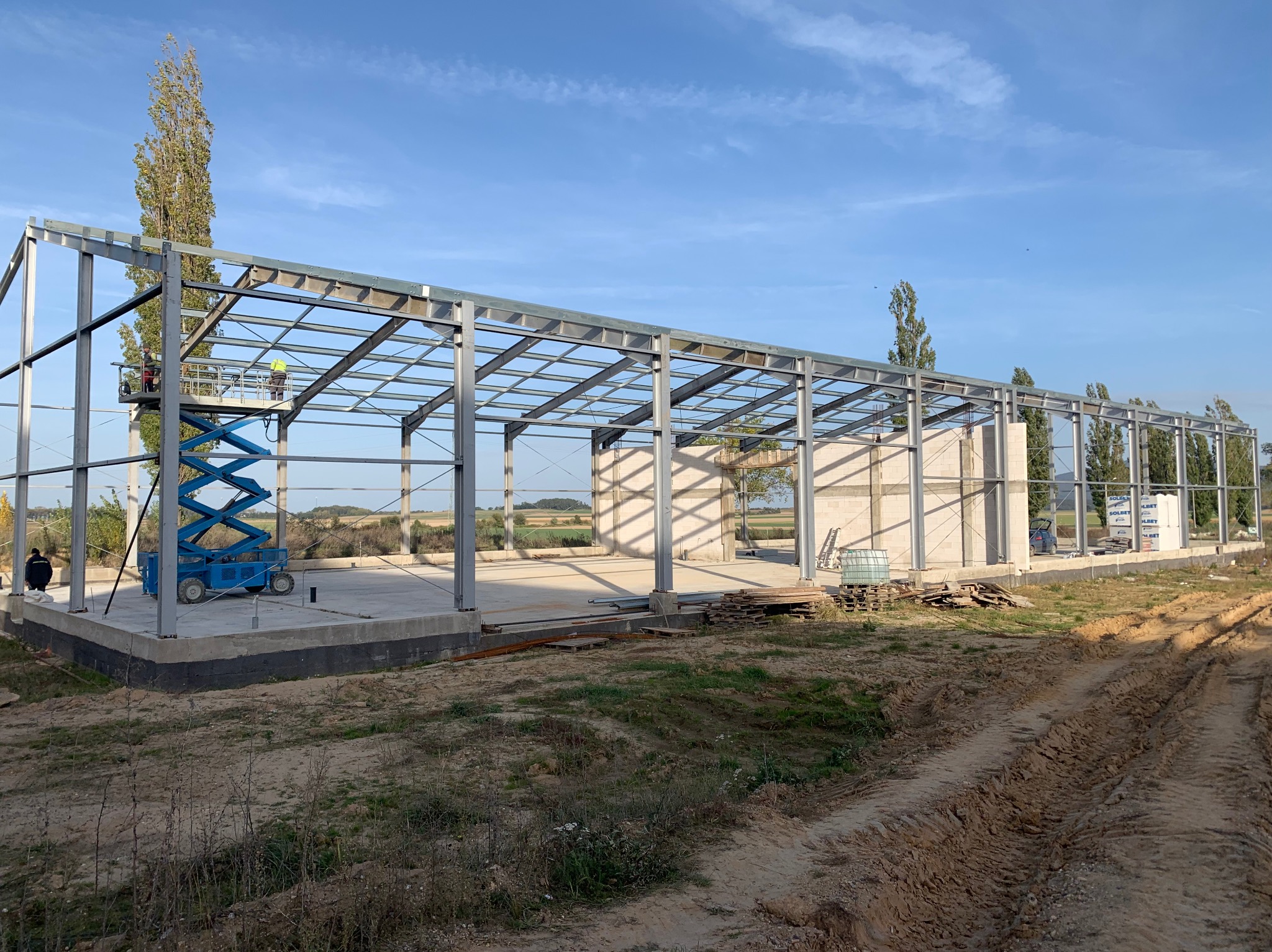 Konstrukcja stalowa hali w trakcie budowy, widoczny podnośnik koszowy z pracownikiem, fundamenty i częściowo wzniesione ściany z betonu komórkowego.