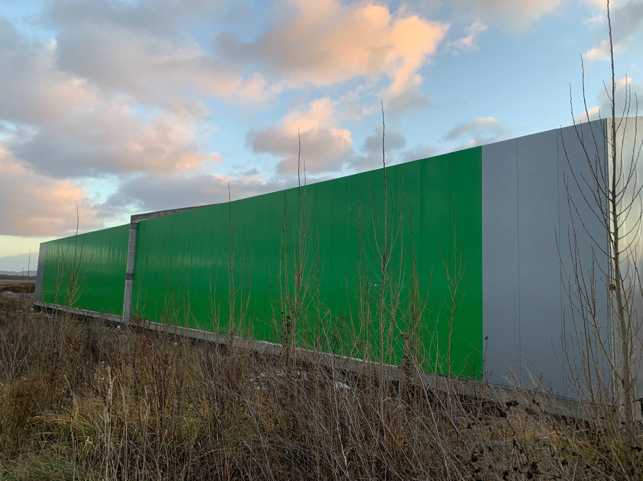 Zewnętrzny widok hali z zielonymi i szarymi panelami, widzianej z poziomu gruntu przez wyschniętą trawę. Częściowo zachmurzone niebo w tle.