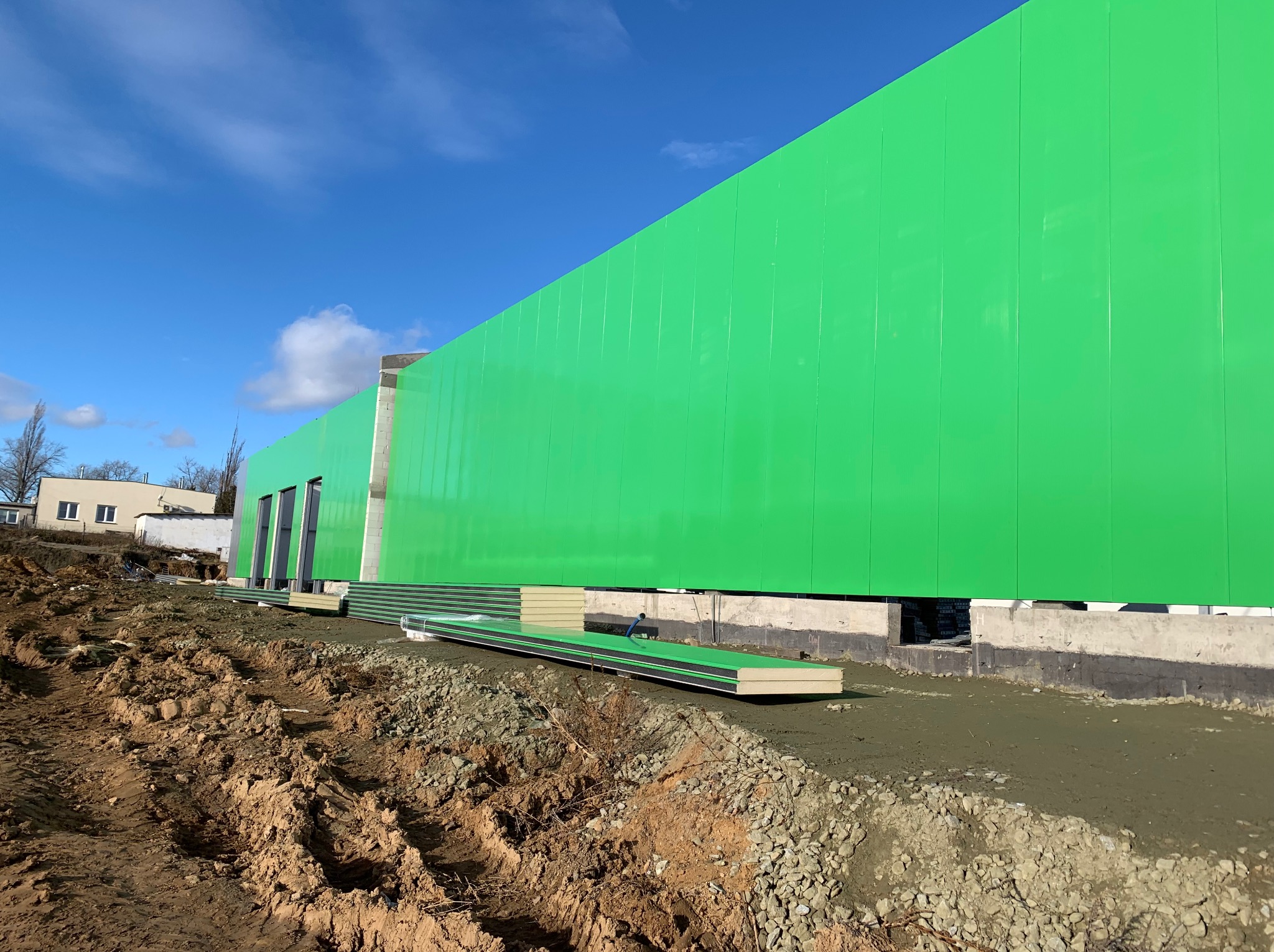 Wznoszenie hali: fragment ściany z paneli w jaskrawej zieleni, widoczne fundamenty i przygotowane płyty warstwowe. Błękitne niebo w tle.