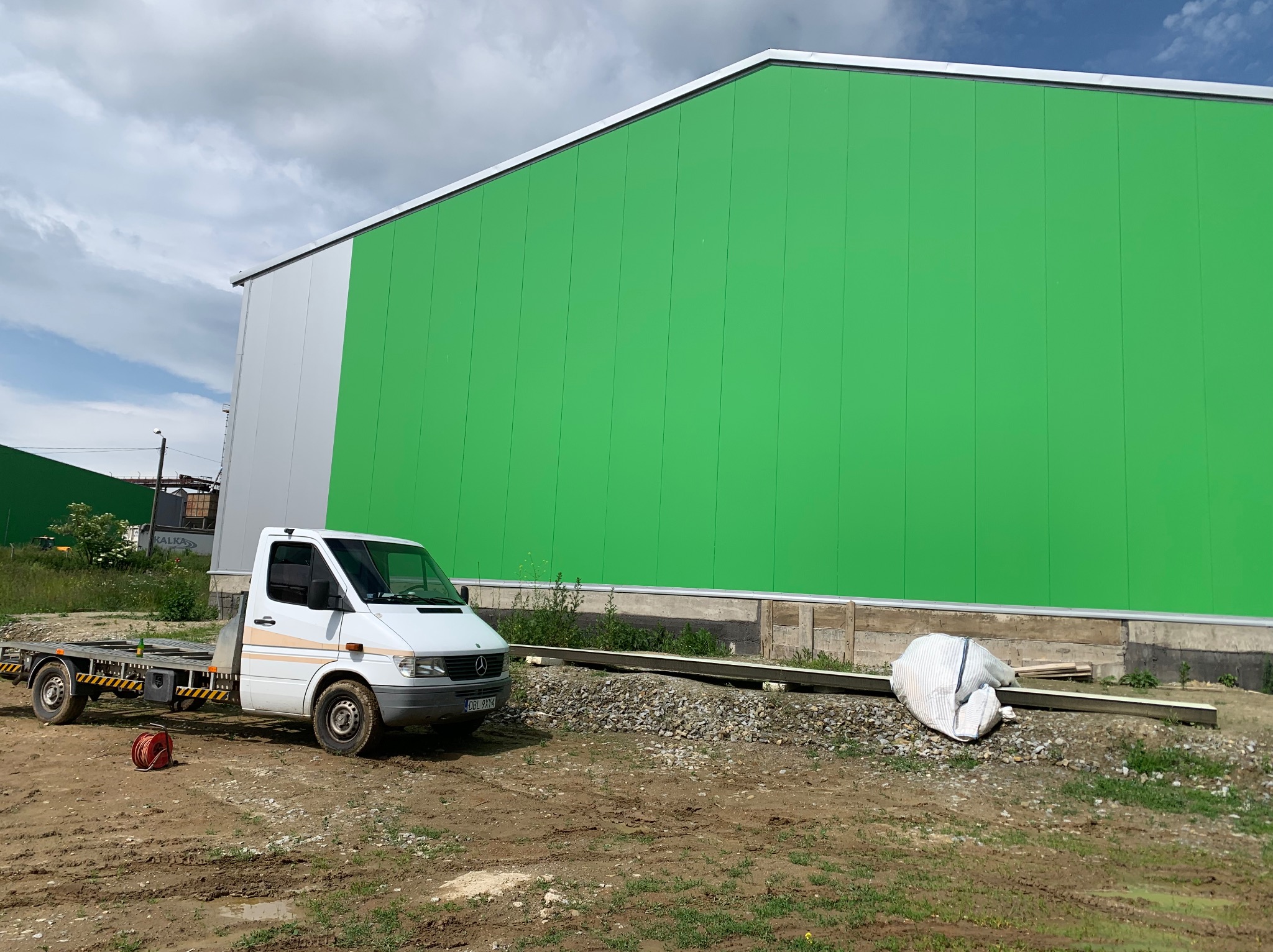Nowoczesna hala w zielono-srebrnej kolorystyce, z transportem na placu budowy. Widoczne elementy konstrukcyjne i teren przygotowany pod dalsze prace.