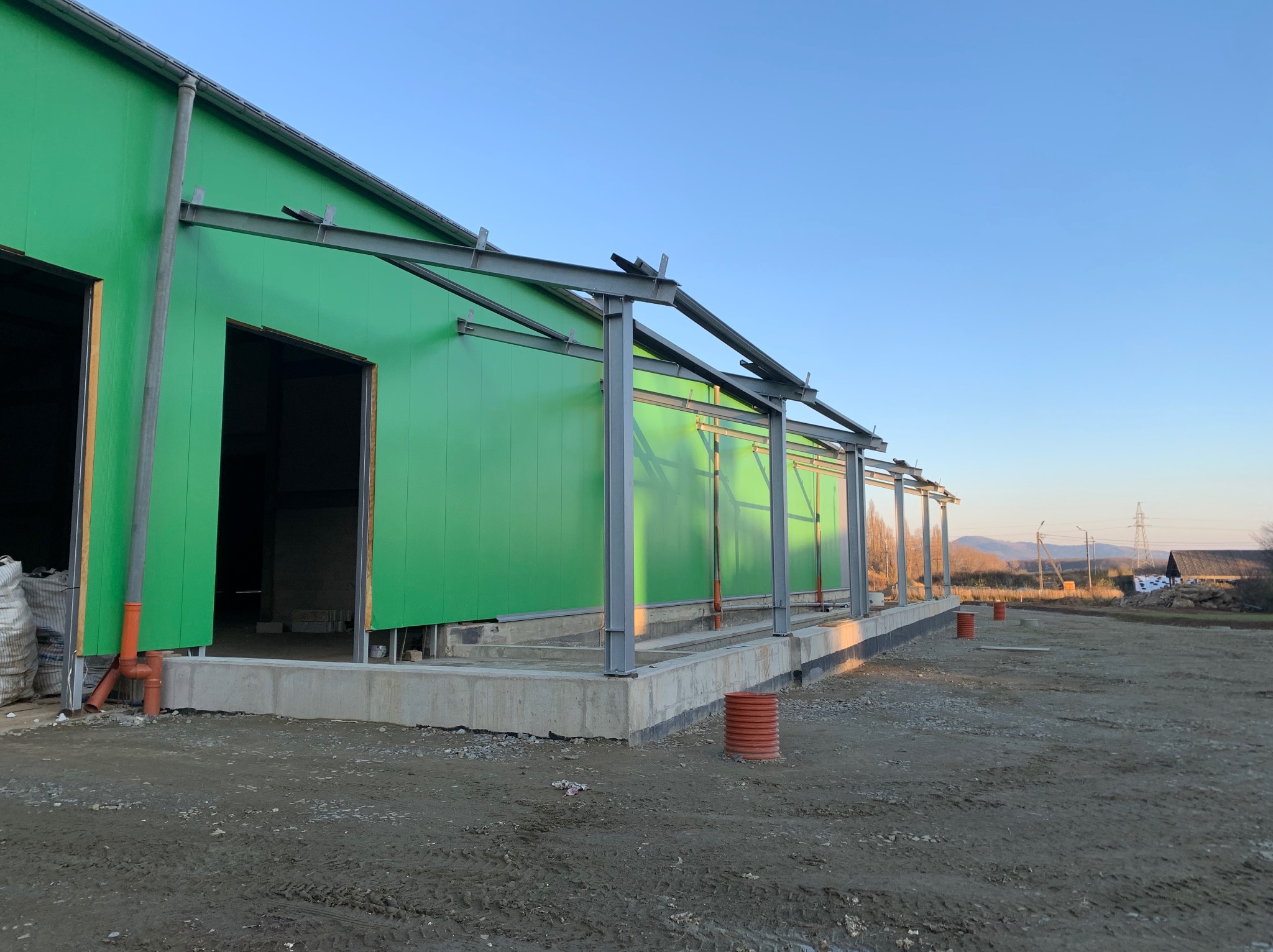 Zielona hala w trakcie budowy, widoczne stalowe elementy konstrukcyjne i fundamenty. W tle krajobraz z drzewami i wzgórzami, błękitne niebo.