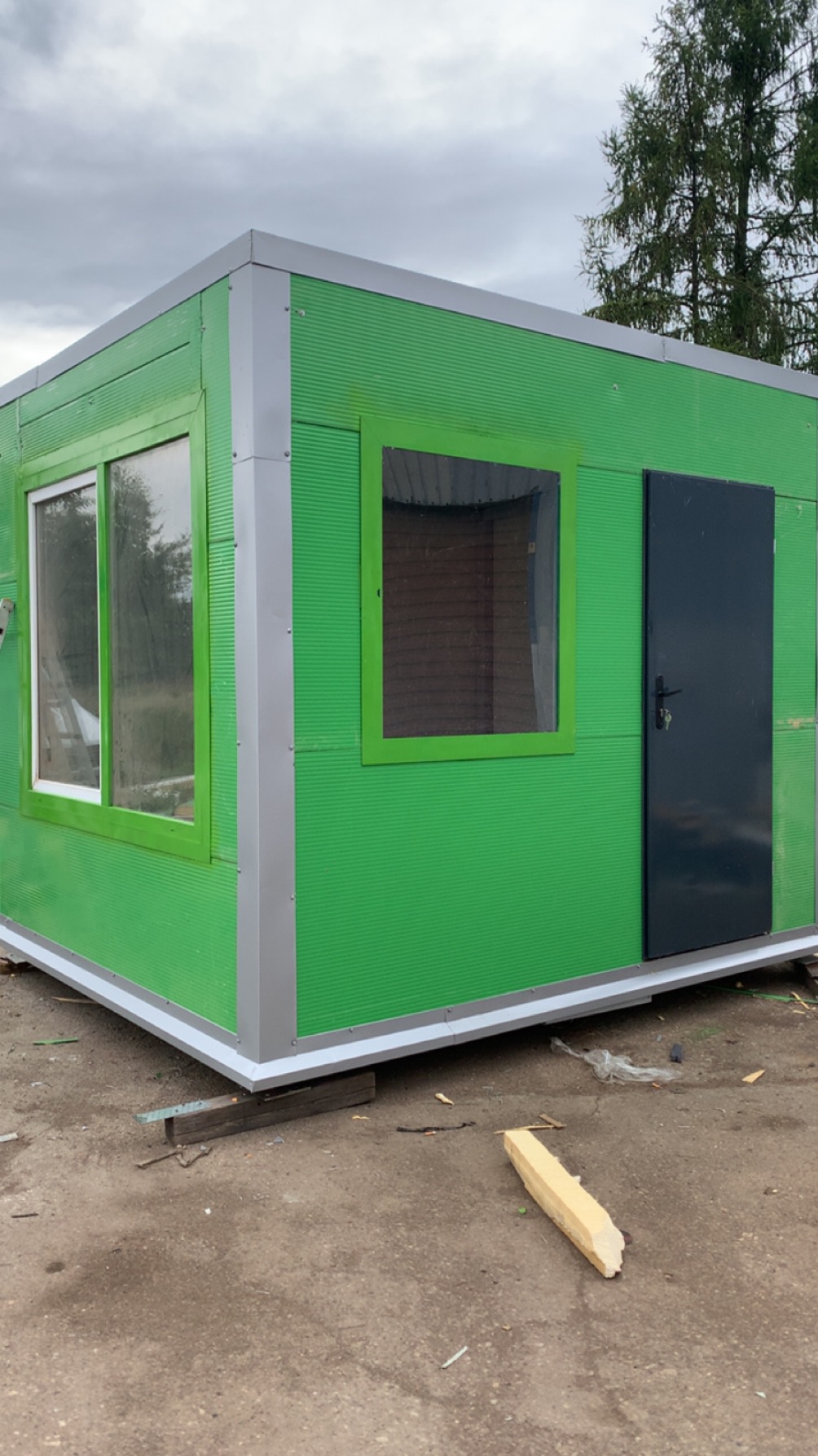 Zielony, modułowy kontener biurowy z oknami i czarnymi drzwiami, ustawiony na placu budowy. Widoczne deski podtrzymujące konstrukcję i fragment drzewa w tle.