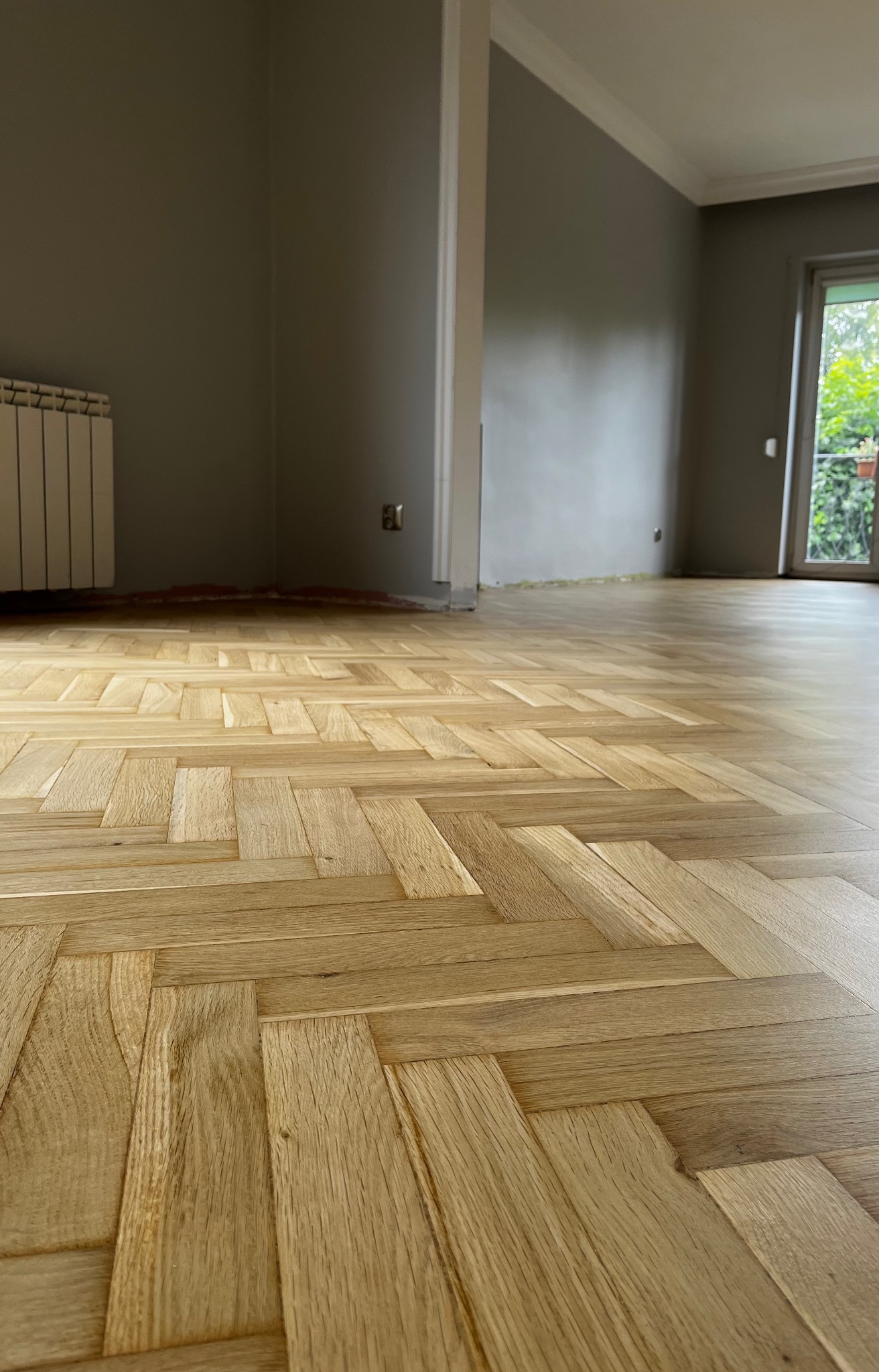 Jasny parkiet ułożony w jodełkę francuską w przestronnym pokoju z szarymi ścianami i grzejnikiem. Widok na wykończoną podłogę po cyklinowaniu i lakierowaniu.