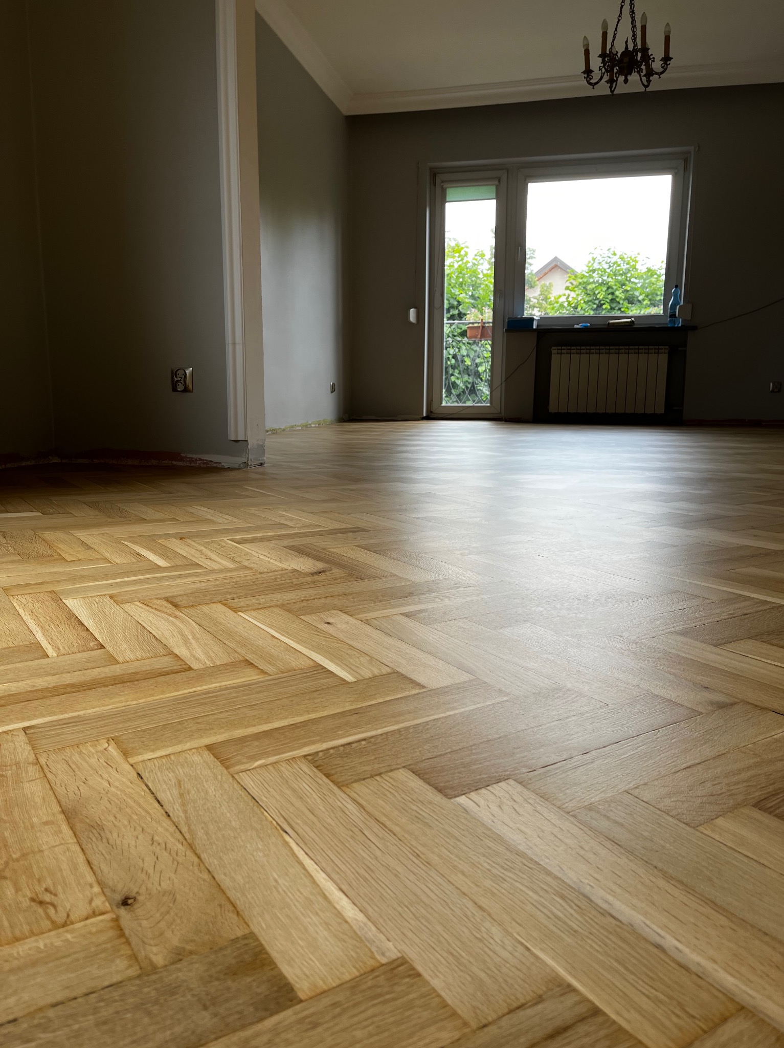 Jasny parkiet ułożony w jodełkę w przestronnym pokoju z oknem i grzejnikiem. Widoczne ślady prac wykończeniowych przy ścianie. Żyrandol w tle.