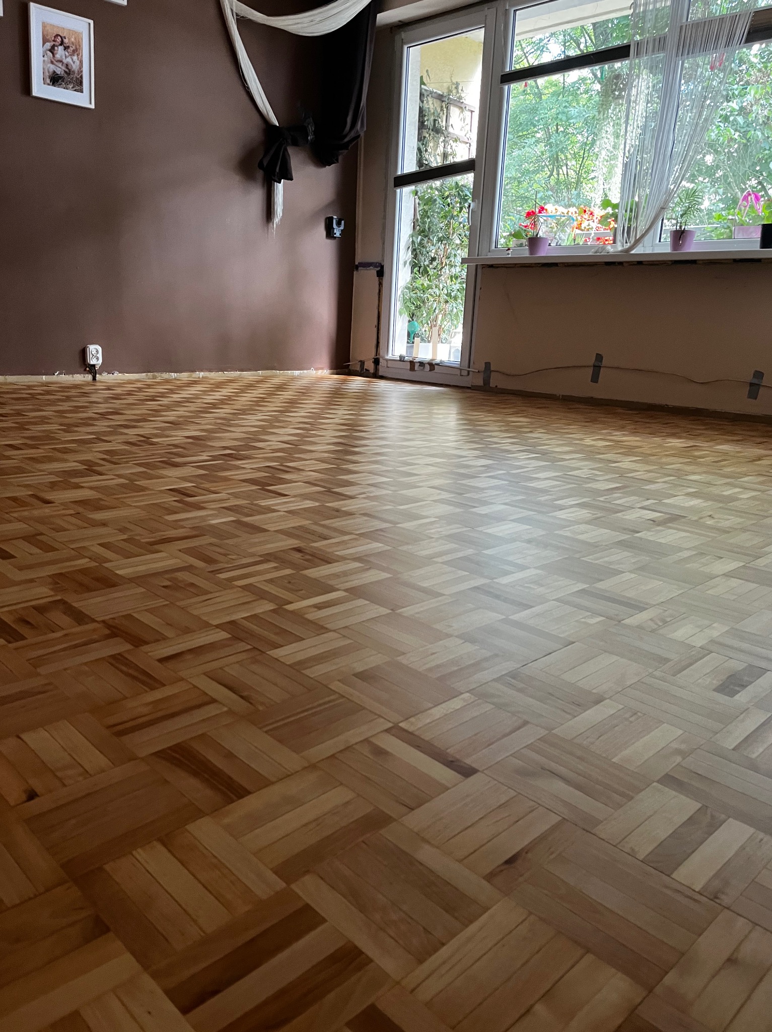 Parkiet ułożony w jodełkę na brązowo-beżowej podłodze, widok z góry. W tle brązowa ściana, okno z kwiatami i dekoracje.