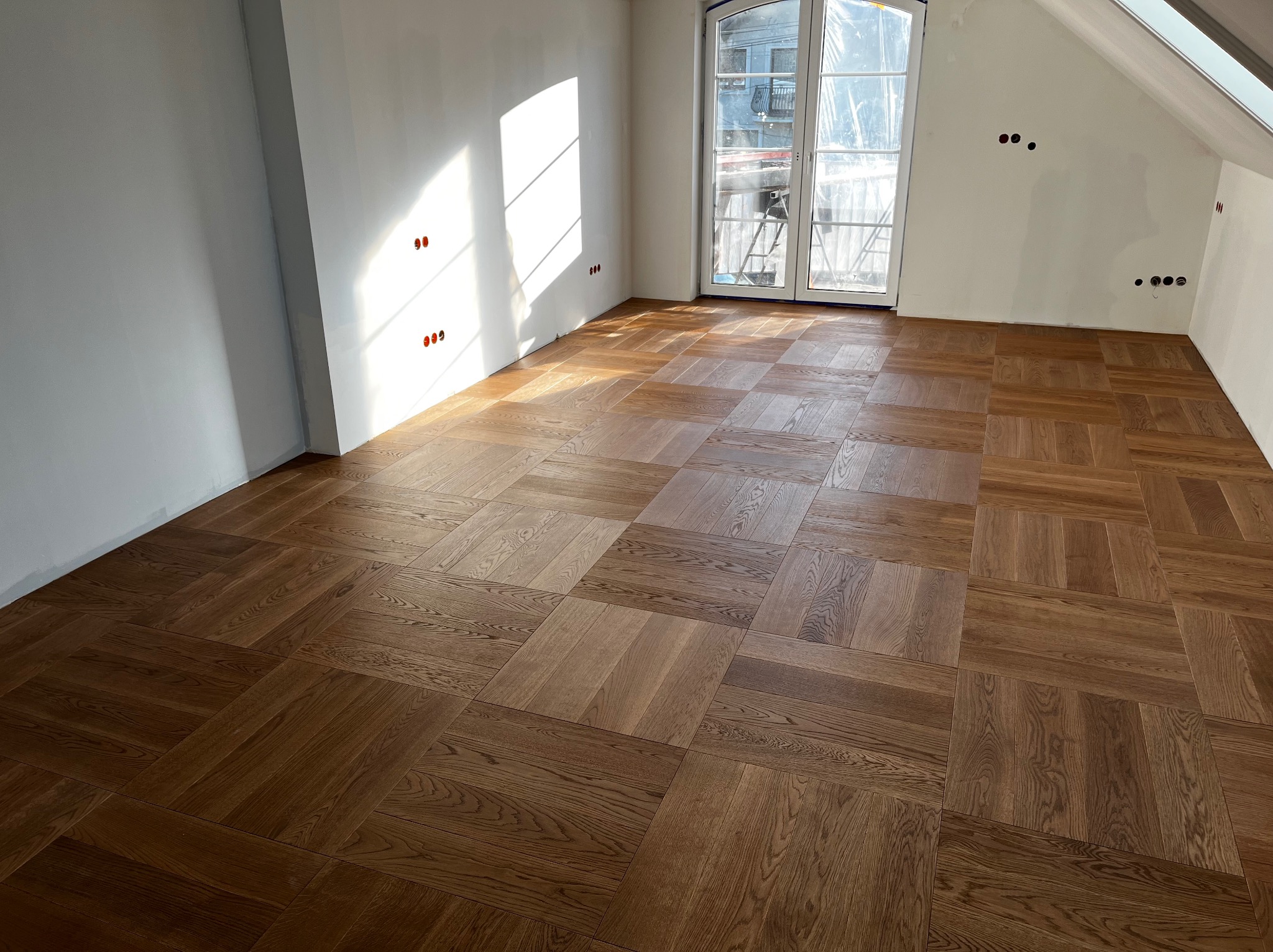 Ułożony parkiet w jodełkę francuską w jasnym pokoju z oknem balkonowym. Widoczne gniazdka elektryczne na ścianie. Parkiet z wyraźnym rysunkiem drewna.
