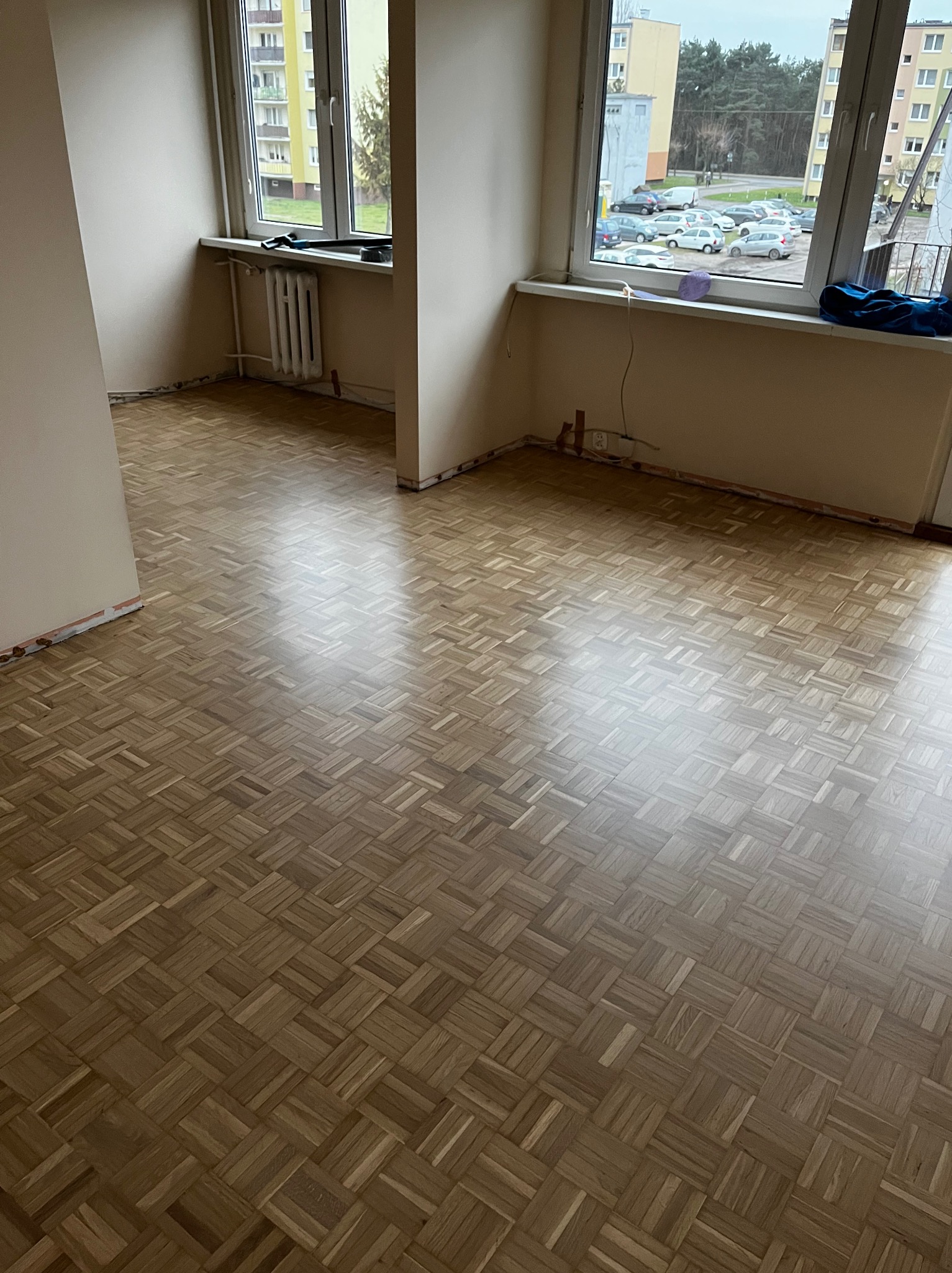 Jasny parkiet ułożony w kwadraty w pustym pokoju z oknami. Widoczne ślady po listwach przypodłogowych i grzejnik. Za oknem blok mieszkalny i drzewa.