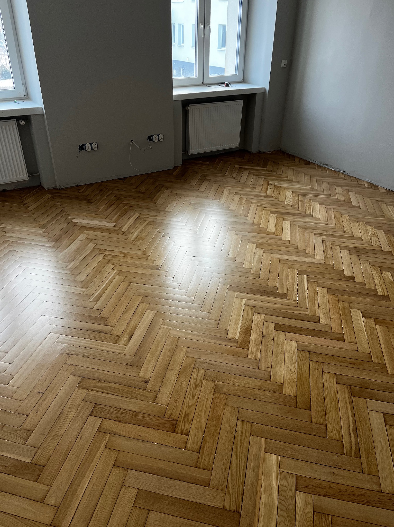 Parkiet ułożony w jodełkę w jasnym pomieszczeniu z oknami i grzejnikami. Widoczne gniazdka elektryczne na szarej ścianie. Świeżo położona podłoga z drewna.