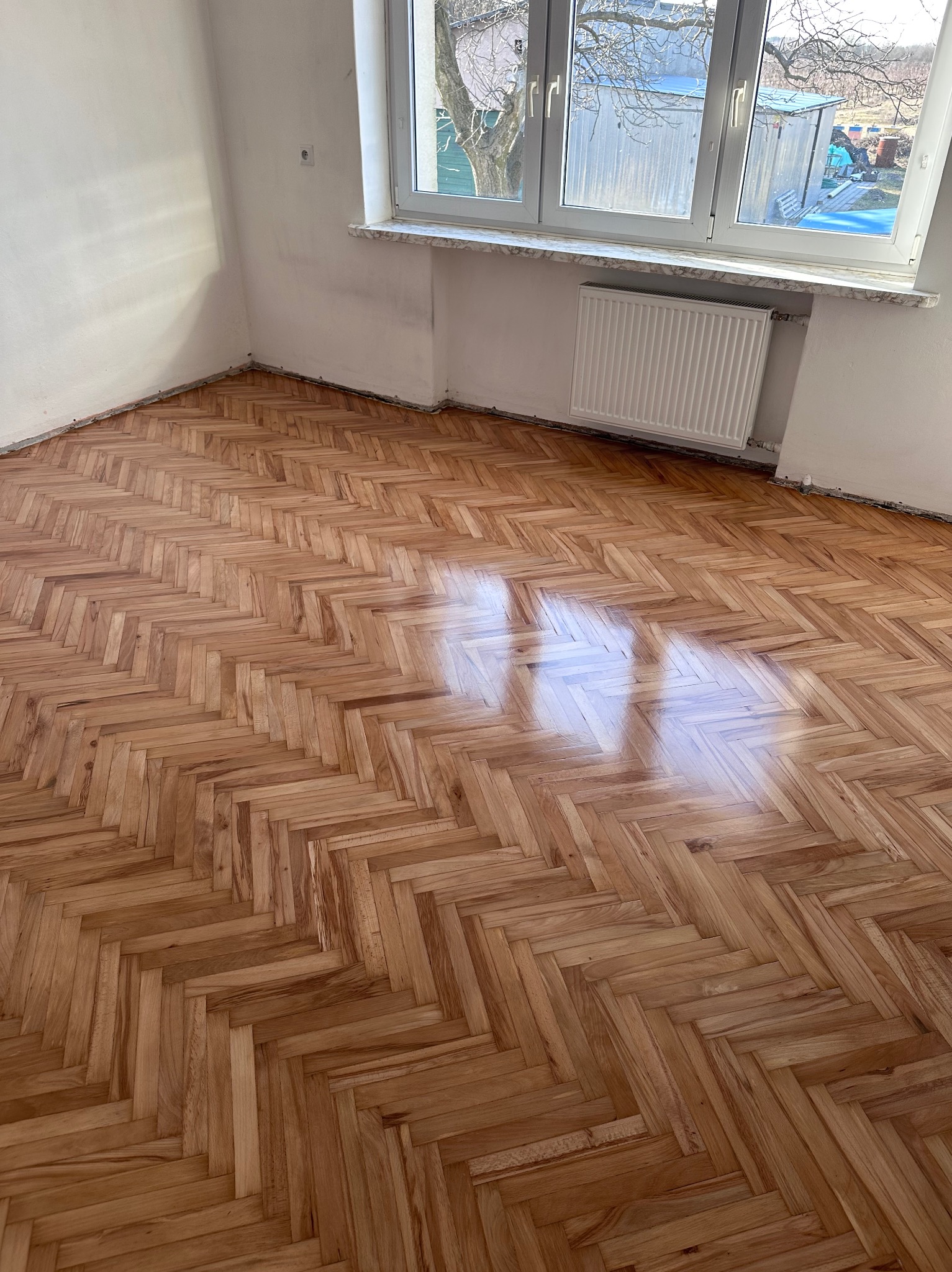 Parkiet ułożony w jodełkę, świeżo po renowacji, z widocznymi refleksami światła. Białe ściany i okno z widokiem na drzewo w tle.