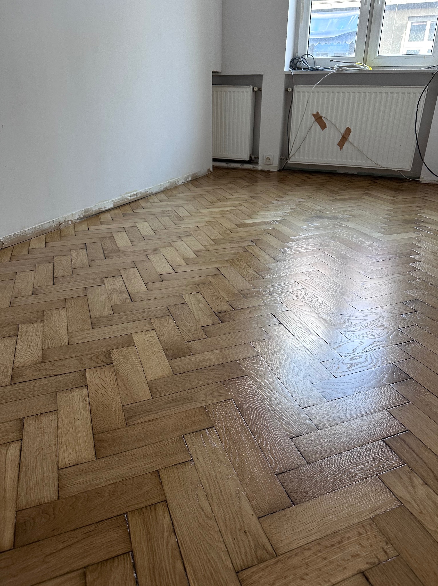 Ukończona podłoga z parkietu ułożonego w jodełkę, po renowacji. Widoczne ślady po taśmie malarskiej na ścianie i grzejniki. Jasne wnętrze z naturalnym oświetleniem.