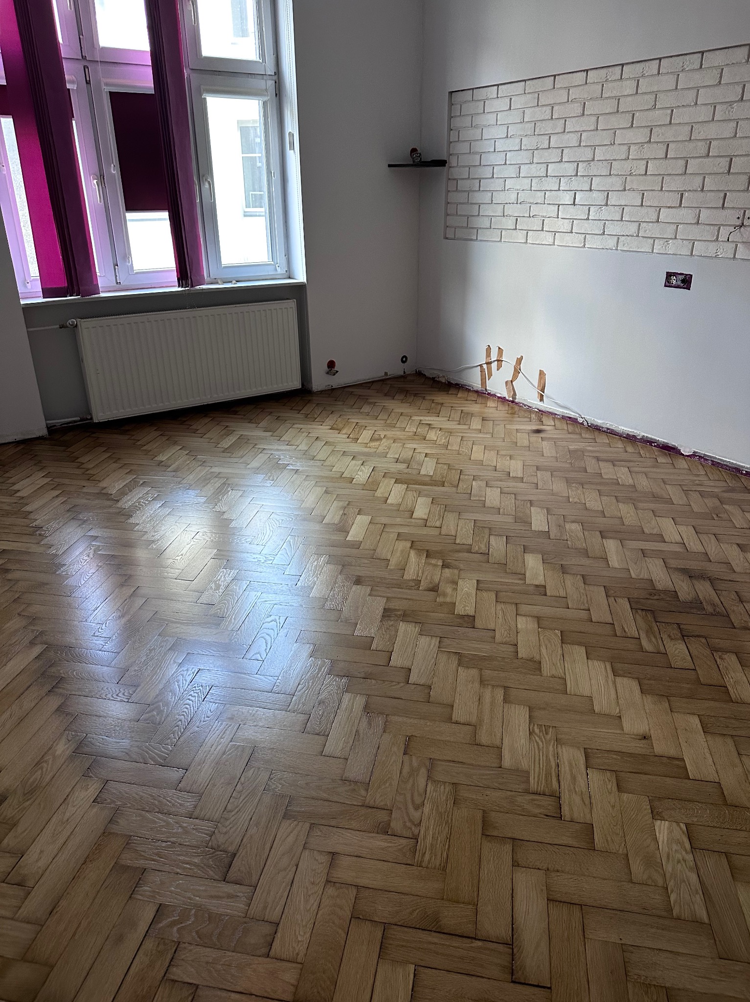 Parkiet ułożony w jodełkę w pustym pokoju z oknem i grzejnikiem. Ściana z cegieł i wystające kable. Widoczne zabezpieczenia przy podłodze.