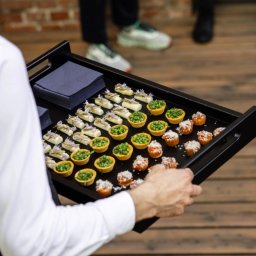 Concordia Design Catering - Elegancka czarna taca z różnorodnymi, apetycznymi przekąskami cateringowymi, trzymana przez kelnera w białej koszuli. Widoczne tartaletki z groszkiem i mini kanapeczki.