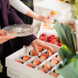 Concordia Design Catering - Eleganckie słodkości w pudełkach: kobieta w bordowej marynarce sięga po deser w kształcie brzoskwini. W tle inne desery na talerzach, tworząc wykwintną kompozycję.