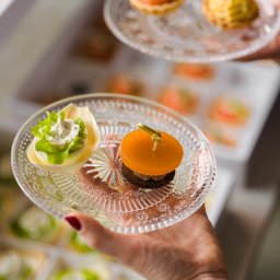 Concordia Design Catering - Eleganckie, przezroczyste talerzyki z wykwintnymi przekąskami cateringowymi, trzymane w dłoniach z widocznym zegarkiem, na tle pudełek z identycznymi przystawkami.