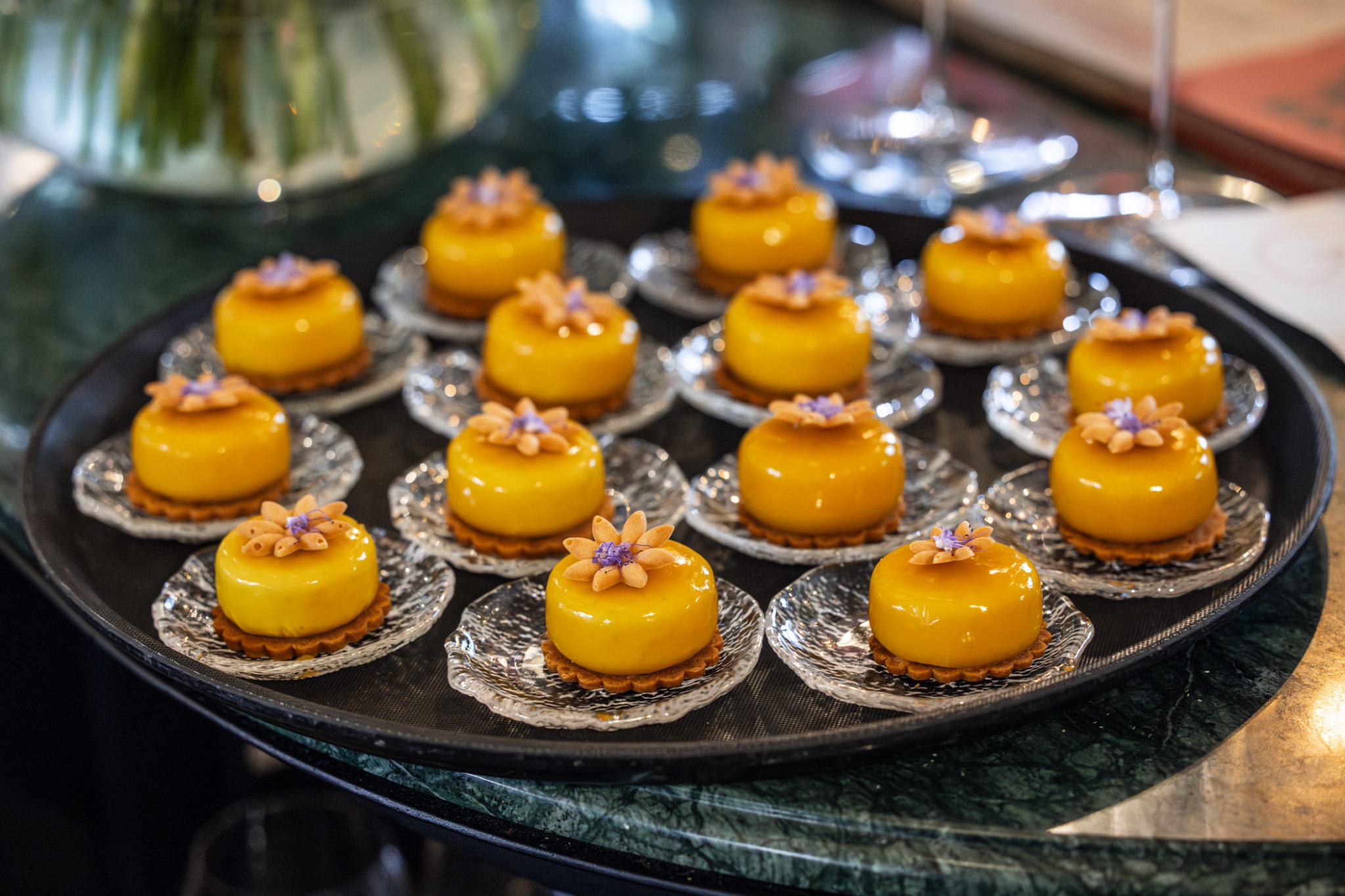 Eleganckie, żółte mini-desery z kwiatami na kruchym spodzie, ułożone na szklanych talerzykach na czarnej tacy. W tle rozmyte szkło i marmurowy blat.
