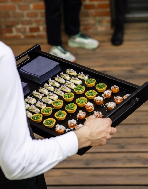 Elegancka czarna taca z różnorodnymi, apetycznymi przekąskami cateringowymi, trzymana przez kelnera w białej koszuli. Widoczne tartaletki z groszkiem i mini kanapeczki.