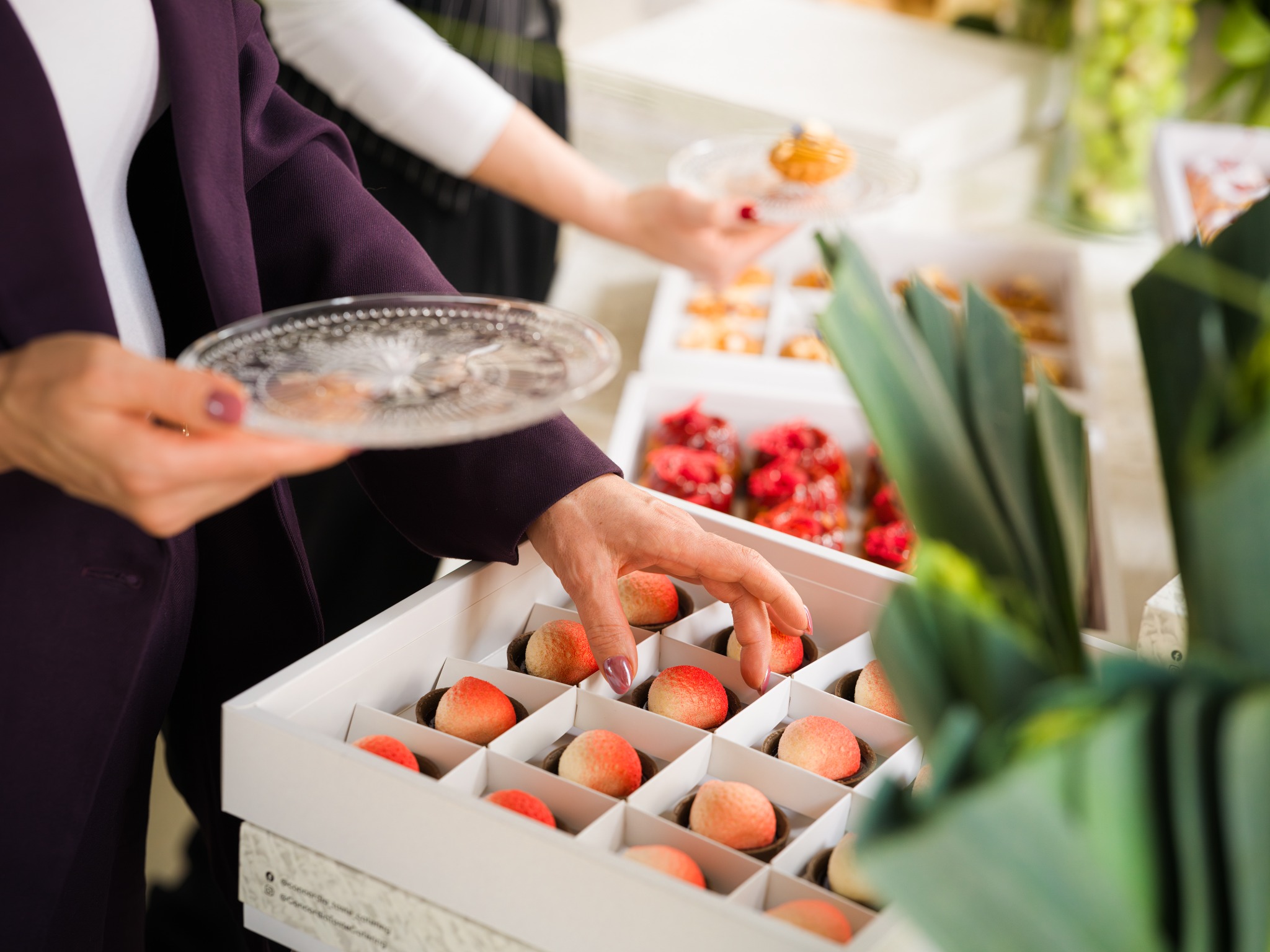 Eleganckie słodkości w pudełkach: kobieta w bordowej marynarce sięga po deser w kształcie brzoskwini. W tle inne desery na talerzach, tworząc wykwintną kompozycję.