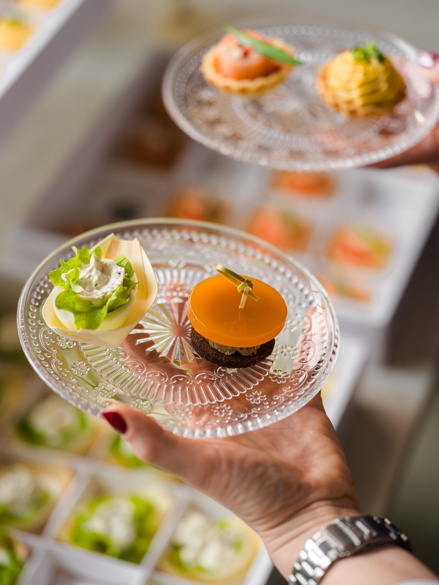 Eleganckie, przezroczyste talerzyki z wykwintnymi przekąskami cateringowymi, trzymane w dłoniach z widocznym zegarkiem, na tle pudełek z identycznymi przystawkami.