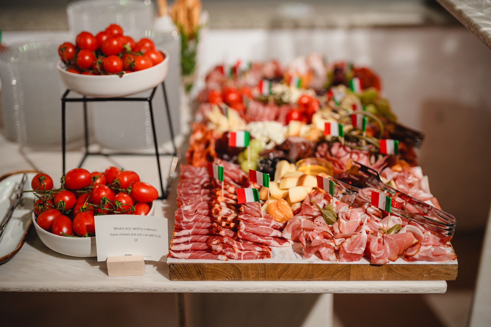 Stół cateringowy z włoskimi serami, wędlinami i owocami, udekorowany flagami Włoch. Dwie misy z pomidorami koktajlowymi. Bufet w neutralnej kolorystyce.