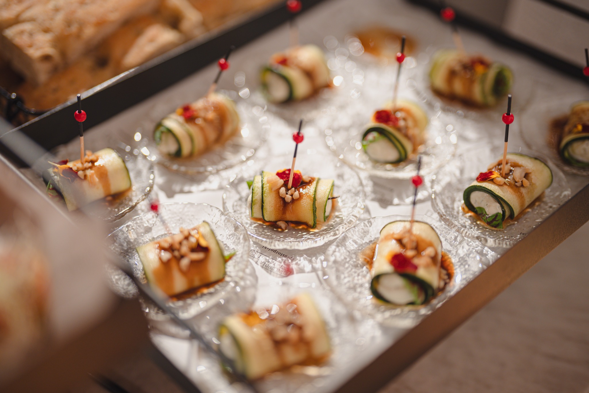 Ekskluzywne roladki z cukinii z kremowym nadzieniem, udekorowane orzechami i sosem, podane na małych, szklanych talerzykach. Elegancki catering na przyjęcie.