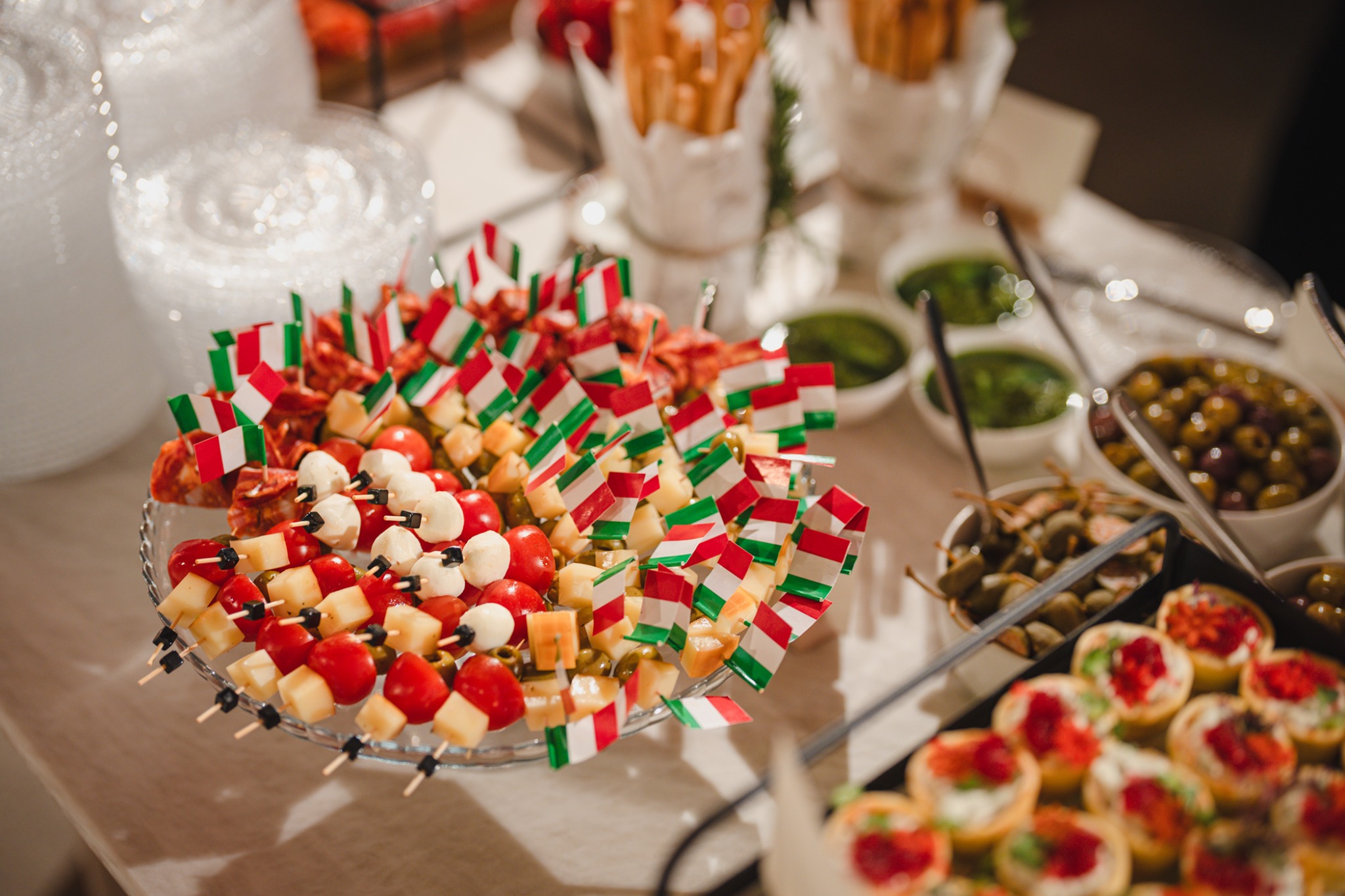 Elegancki bufet z przekąskami na przyjęciu: koreczki z flagami Włoch, oliwki, grissini i mini tartaletki. Jasne, naturalne światło.