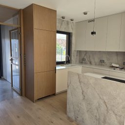 PRESTIGE DECOR ROBERT MOCZULSKI - Nowoczesna kuchnia z wyspą wykończoną marmurem, drewnianymi szafkami i parkietem. Minimalistyczne oświetlenie punktowe i okno z widokiem na zieleń.