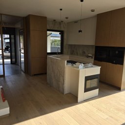 PRESTIGE DECOR ROBERT MOCZULSKI - Nowoczesna kuchnia z wyspą i drewnianymi frontami, jasna podłoga, minimalistyczne oświetlenie. Widok na okno i fragment otoczenia zewnętrznego.