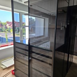 PRESTIGE DECOR ROBERT MOCZULSKI - Montaż szafy wnękowej z ciemnymi, przesuwnymi drzwiami i drewnianym wnętrzem. Widoczne narzędzia i elementy montażowe. Duże okno z widokiem na domy w tle.