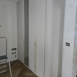 PRESTIGE DECOR ROBERT MOCZULSKI - Biała szafa wnękowa z przeszklonym segmentem i złotymi uchwytami, na parkiecie w jodełkę, widoczna drabina malarska i niedokończone wykończenie ściany.