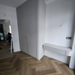 PRESTIGE DECOR ROBERT MOCZULSKI - Jasnoszara szafa z zaokrąglonymi krawędziami i złotymi uchwytami, dopasowana kolorystycznie półka, parkiet w jodełkę. Nowoczesny design.