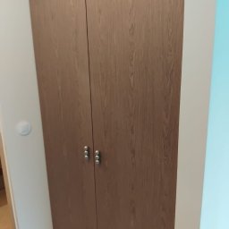 PRESTIGE DECOR ROBERT MOCZULSKI - Dwudrzwiowa szafa wnękowa z drewnopodobnymi frontami i srebrnymi uchwytami, umieszczona w rogu pomieszczenia z widocznym fragmentem ściany w kolorze błękitnym.