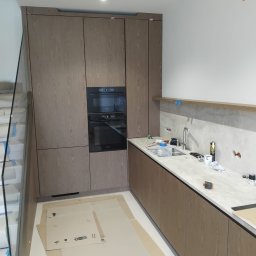 PRESTIGE DECOR ROBERT MOCZULSKI - Montaż nowoczesnej kuchni z drewnianymi frontami i wbudowanym piekarnikiem. Widoczne zabezpieczenia podłogi kartonem. Ujęcie z perspektywy schodów.
