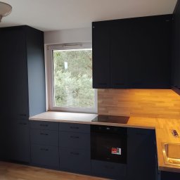 Prestige Decor - Nowoczesna kuchnia w odcieniach granatu z drewnianym blatem i oświetleniem podszafkowym. Widok na okno z zielenią w tle. Minimalistyczny design, zabudowa pod sam sufit.