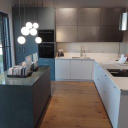 Prestige Decor - Nowoczesna kuchnia z wyspą i drewnianą podłogą. Widoczne próbki materiałów na blacie i mężczyzna przeglądający dokumenty. Minimalistyczny design, oświetlenie punktowe.