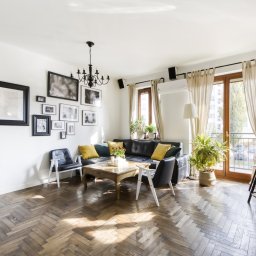 Marek Bieńko - Jasne wnętrze salonu z parkietem w jodełkę, czarną sofą z żółtymi poduszkami, galerią obrazów na ścianie i wyjściem na balkon. Widok na przestronne, urządzone mieszkanie.