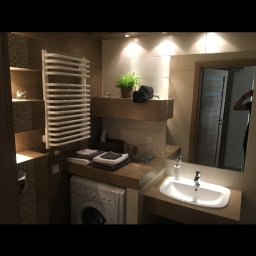 Sławomir Grządziel - Jasna łazienka z pralką pod blatem, grzejnikiem drabinkowym i dużym lustrem. Umywalka nablatowa, beżowe płytki i drewniane akcenty tworzą przytulne wnętrze.