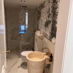 Sławomir Grządziel - Elegancka łazienka z marmurowymi płytkami, wolnostojącą umywalką z trawertynu i dekoracyjną tapetą w stylu vintage. Widoczna kabina prysznicowa i złote akcenty.