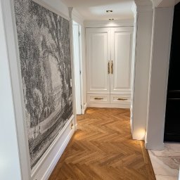 Sławomir Grządziel - Elegancki korytarz z parkietem w jodełkę, białą zabudową i dekoracyjną grafiką na ścianie. Oświetlenie punktowe podkreśla detale wykończenia.