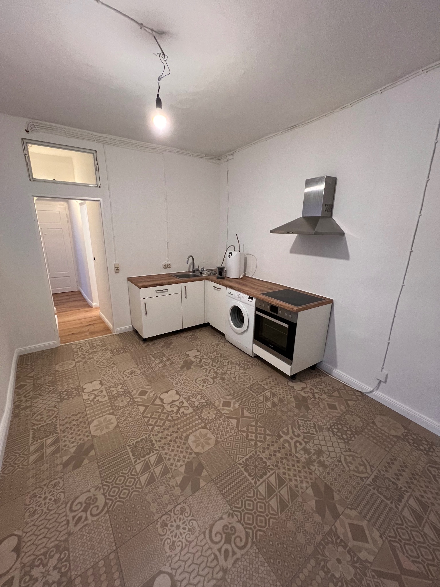 Wykończona kuchnia z wzorzystą podłogą, białe szafki z drewnianym blatem, pralka i piekarnik w zabudowie. Widok na sąsiednie pomieszczenie.