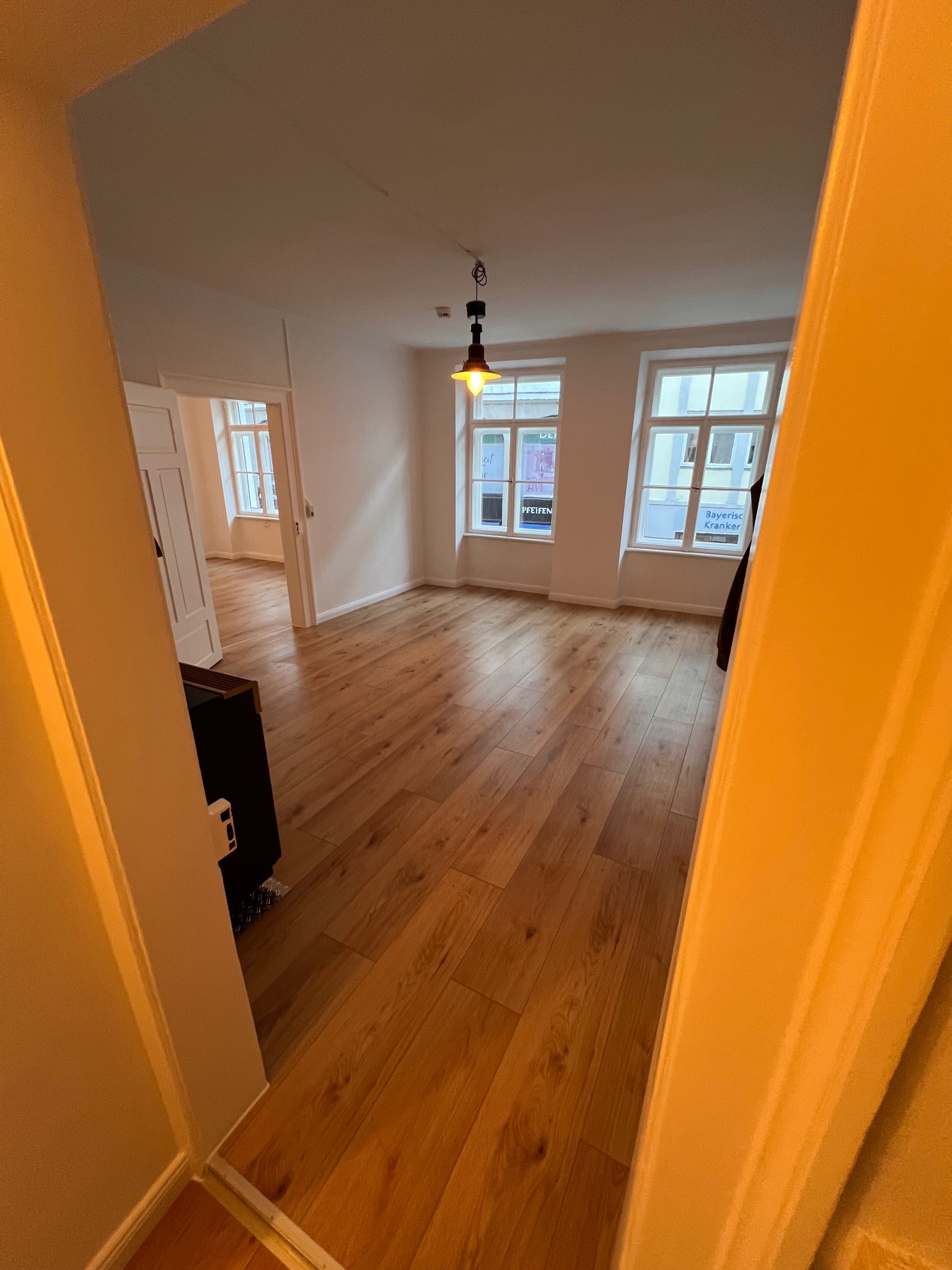 Jasne, przestronne wnętrze z drewnianą podłogą, widoczne zza framugi drzwi. Ściany białe, duże okna wpuszczają światło. Minimalistyczna lampa sufitowa.