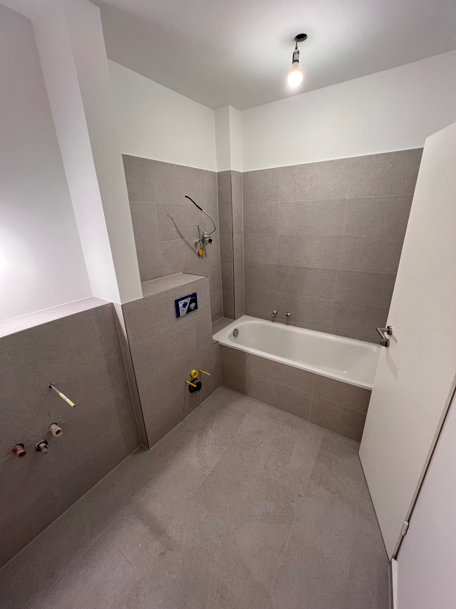 Wykończona łazienka z beżowymi płytkami, białą wanną i częściowo widoczną białą ścianą. Widoczne przyłącza wodne i odpływowe. Minimalistyczny design, surowy stan.