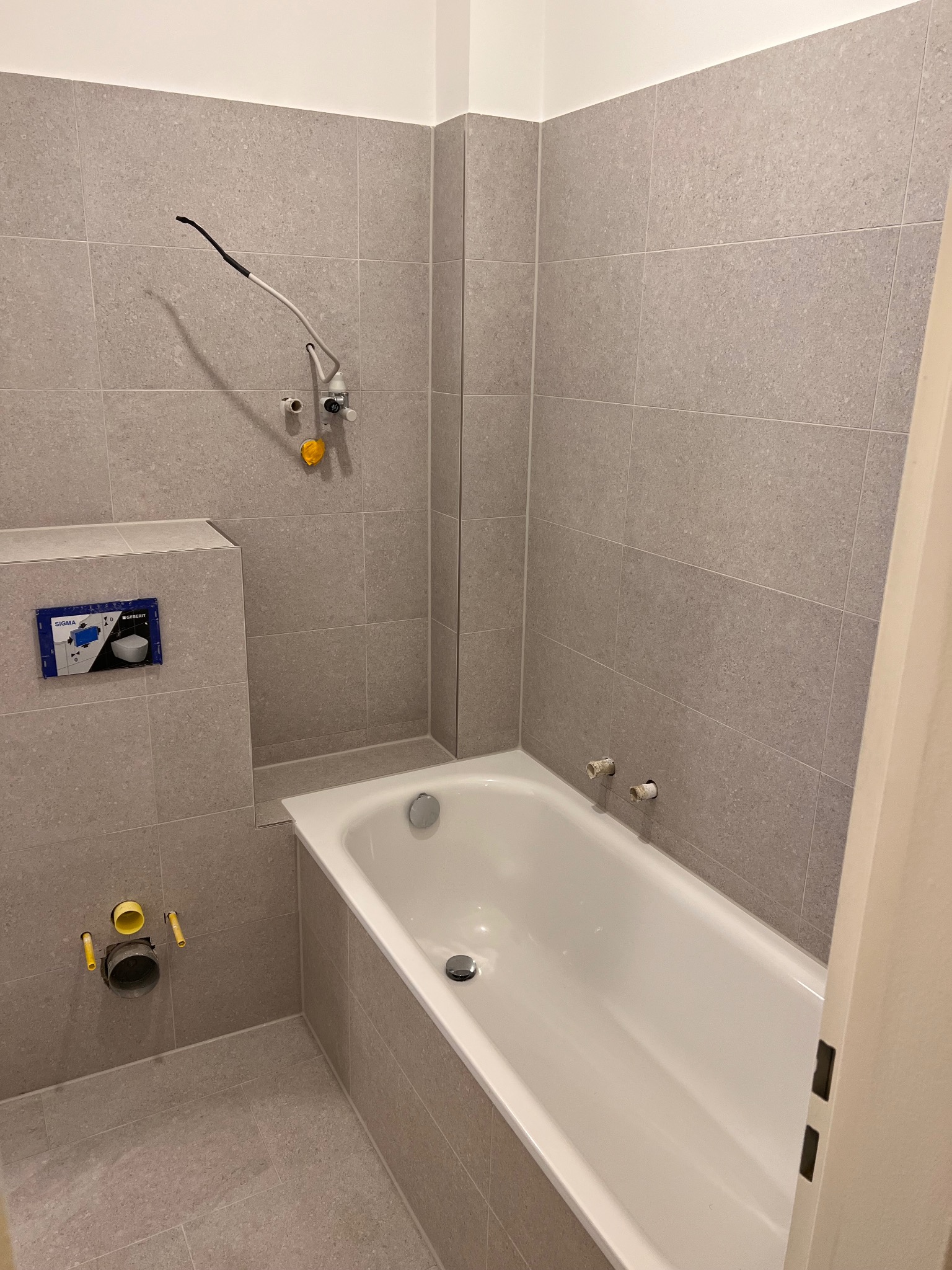 Szara łazienka w trakcie wykańczania: wanna, płytki, instalacja sanitarna z wystającymi rurami i elementami montażowymi. Widok na fragment wnętrza.