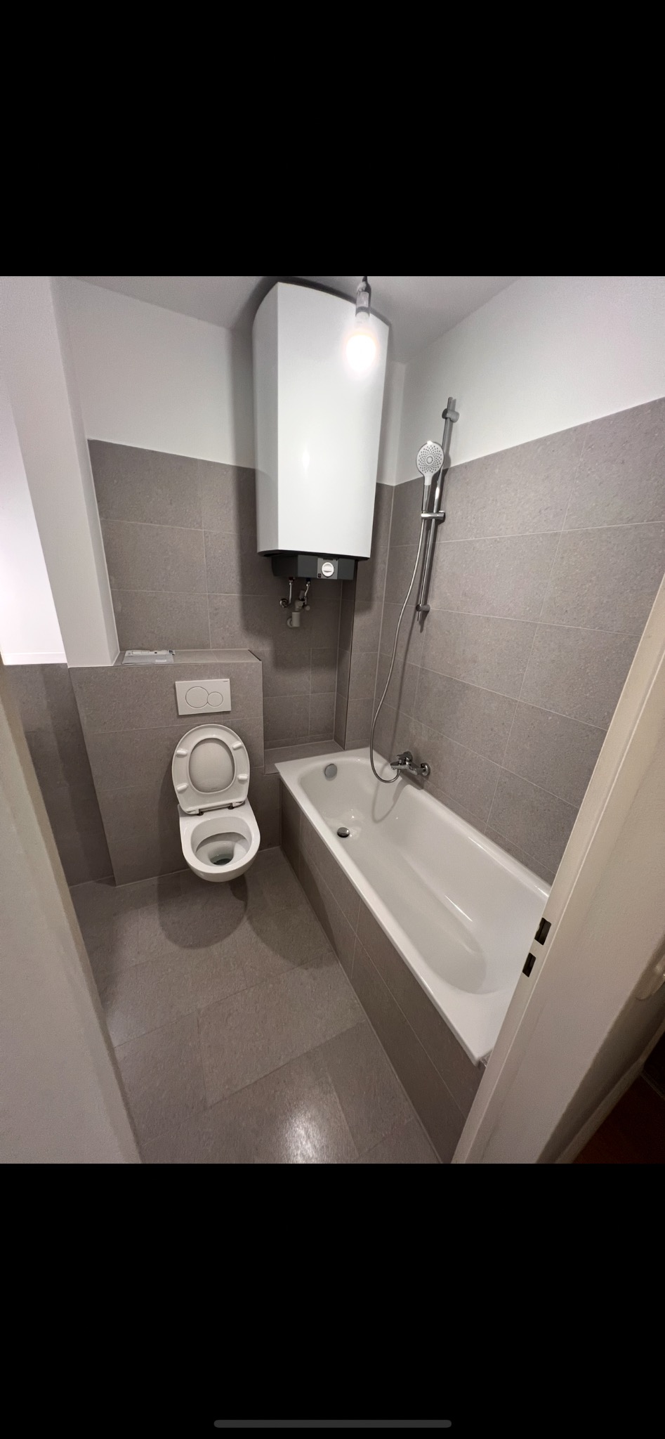 Wykończona łazienka w szarościach: płytki na ścianach i podłodze, biała wanna, toaleta z otwartą klapą i bojler. Minimalistyczny design, widok z perspektywy wejścia.