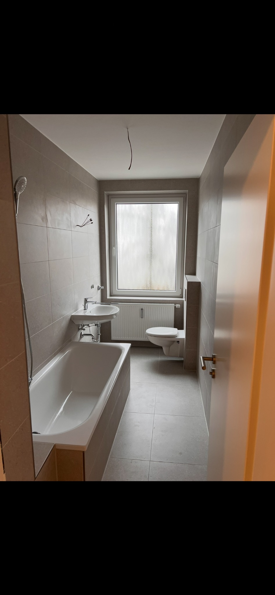 Jasna łazienka z wanną, umywalką i toaletą, wykończona szarymi płytkami. Widoczne okno z grzejnikiem. Uwagę przykuwa kabel zwisający z sufitu.