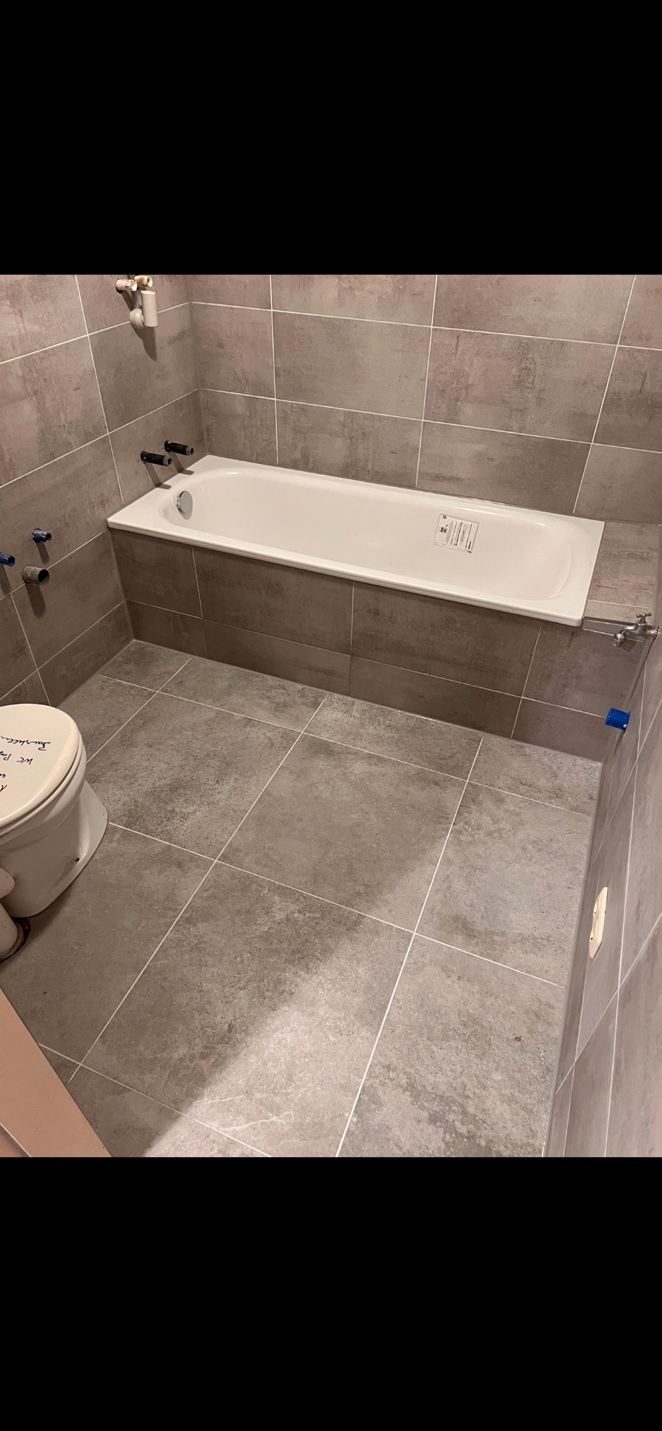 Wykończona łazienka z wanną i szarą, kwadratową glazurą. Widoczne przyłącza wodne i fragment toalety z napisem. Minimalistyczny styl.