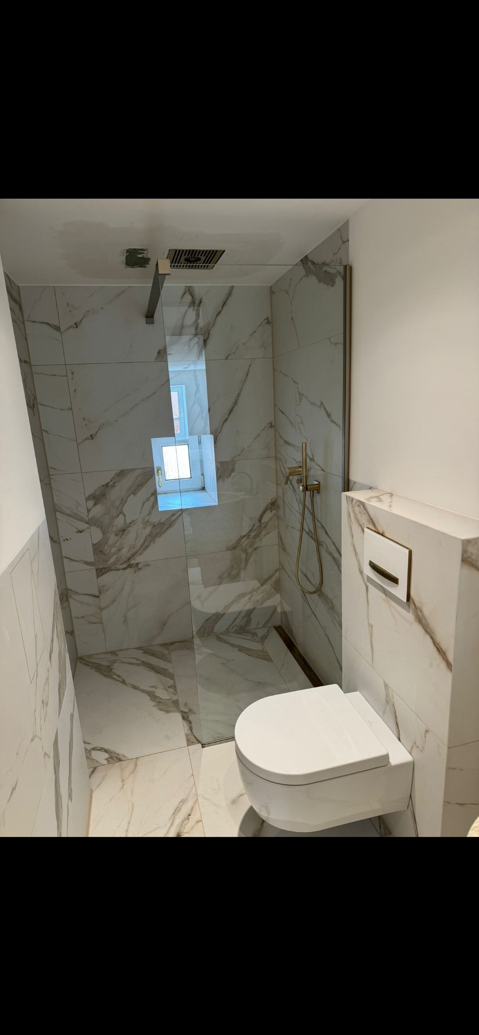 Nowoczesna łazienka z kabiną prysznicową typu walk-in, wykończona białymi płytkami imitującymi marmur. Wisząca toaleta i złota armatura dodają elegancji.