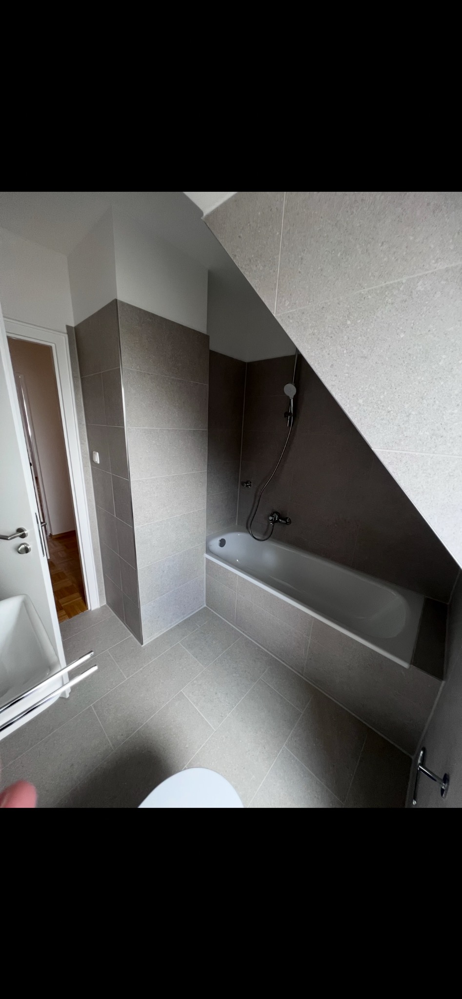 Jasna łazienka na poddaszu z wanną obudowaną płytkami w odcieniach szarości. Umywalka i fragment toalety widoczne w kadrze. Minimalistyczny design.