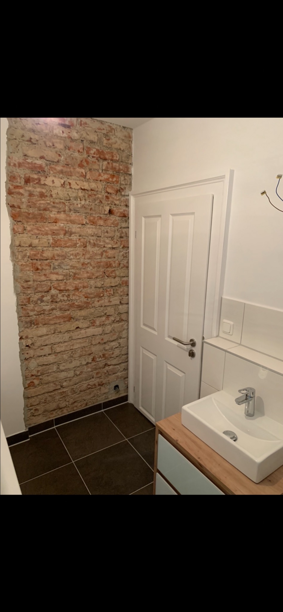 Częściowo wykończona łazienka z odsłoniętą ścianą z cegły, brązowymi płytkami podłogowymi i nowoczesną białą umywalką z szafką z drewnianym blatem.